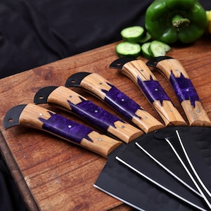 Op de afbeelding: Een set van vijf keukenmessen met houten handvatten en paarse harsinleg. De zwarte messen zijn gebogen. De messen liggen op een houten snijplank. Een groene paprika en gesneden komkommers zijn op de achtergrond te zien.