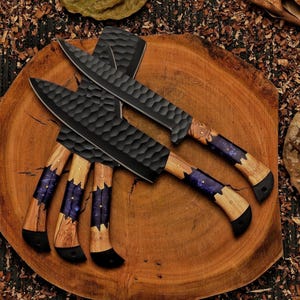 Op de afbeelding: Overzicht van zwarte koksmes met getextureerde messen en houten handvatten. De handvatten hebben een paarse en blauwe harsinleg. De messen liggen op een ronde houten plank, omringd door houtkrullen en bladeren. De messen zijn ongeveer 30 cm lang.