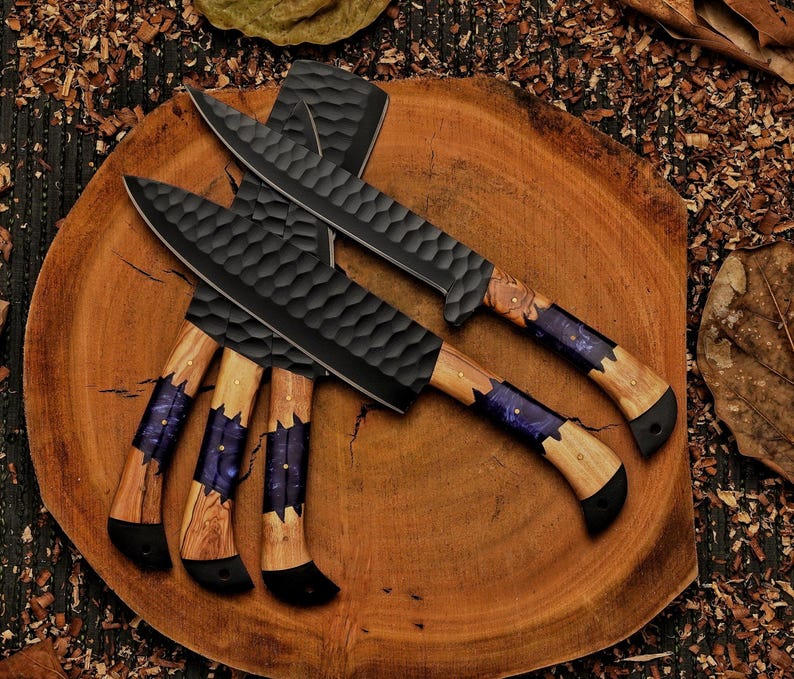 Op de afbeelding: Overzicht van zwarte koksmes met getextureerde messen en houten handvatten. De handvatten hebben een paarse en blauwe harsinleg. De messen liggen op een ronde houten plank, omringd door houtkrullen en bladeren. De messen zijn ongeveer 30 cm lang.