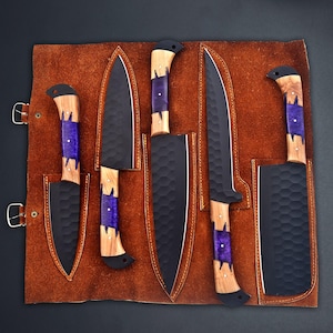 Op de afbeelding: Een set van zes zwarte keukenmessen met houten handvatten en paarse accenten, gepresenteerd in een bruine leren messenrol. De messen hebben een getextureerd oppervlak. De handvatten zijn van hout en paarse hars.