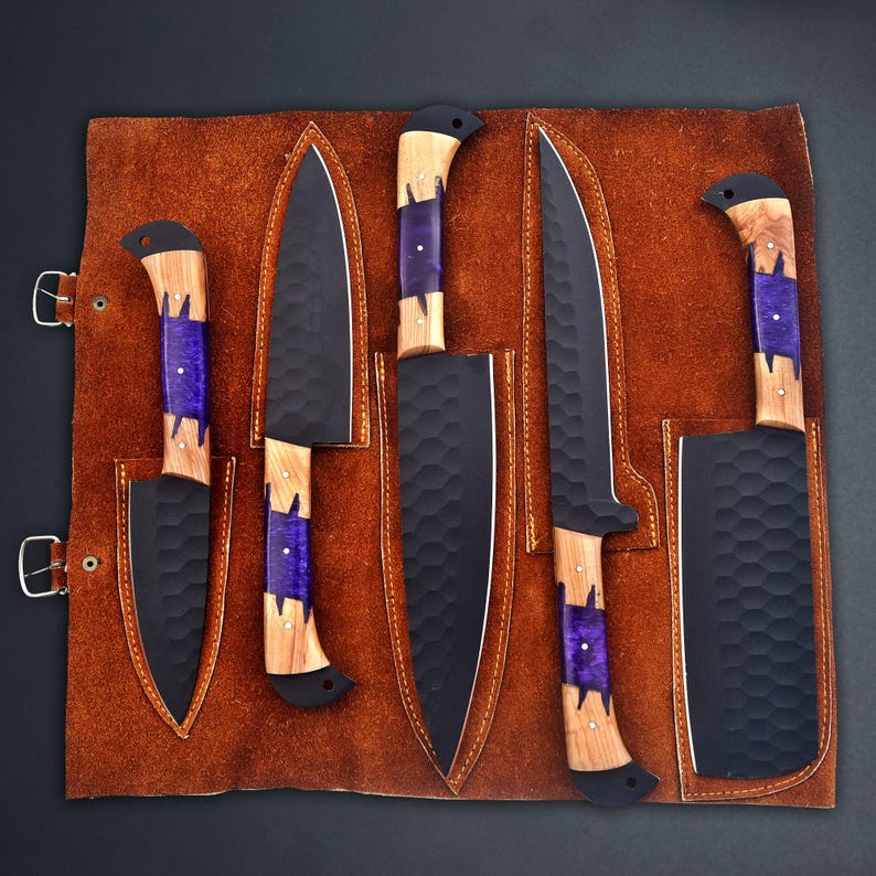 Op de afbeelding: Een set van zes zwarte keukenmessen met houten handvatten en paarse accenten, gepresenteerd in een bruine leren messenrol. De messen hebben een getextureerd oppervlak. De handvatten zijn van hout en paarse hars.