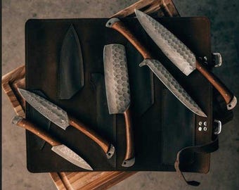 May include: A set of handcrafted knives with wooden handles and patterned blades, displayed on a dark leather knife roll. The set includes various sizes and shapes of knives, including a cleaver. The knife roll is resting on a wooden cutting board.