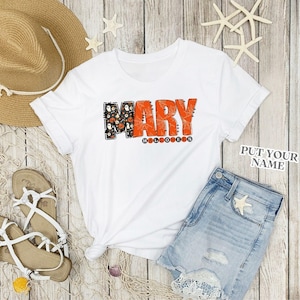 Könnte beinhalten: Weißes T-Shirt mit dem Wort "MARY" in orangefarbenen und schwarzen Halloween-Schriftzügen. Das Shirt wird mit Jeansshorts, Sandalen und einem Strohhut kombiniert. Der Text "PUT YOUR NAME" befindet sich auf einem Aufkleber.
