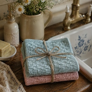 Pode incluir: Uma pilha de dois panos de limpeza quadrados tricotados, um em azul claro e outro em rosa seco, ambos com um padrão de cestaria texturizado. Os panos estão cuidadosamente dobrados e atados com um fio de juta castanho natural fino.