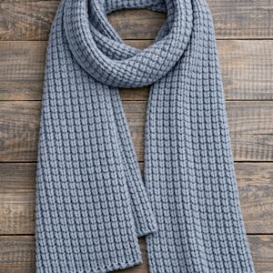 May include: A light blue, ribbed-knit scarf. The scarf is folded in half and draped over a wooden surface. The knit pattern is vertical and creates a textured appearance. The scarf is a solid color and appears to be made of a soft material.