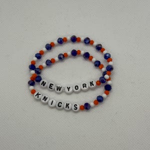 Peut inclure: Deux bracelets élastiques en perles alternant de petites perles orange, blanches et bleu foncé à facettes. Un bracelet présente des perles lettres blanches formant NEW YORK, tandis que l'autre bracelet présente des perles lettres formant KNICKS.