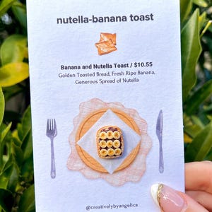 May include: A white card featuring the text nutella-banana toast, Banana and Nutella Toast, and a description of golden toasted bread, fresh ripe banana, and Nutella. It displays a miniature toast with banana slices on a plate, a fork, and a knife.