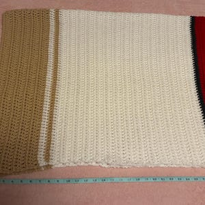 May include: A handmade crochet blanket with a color block design. The blanket features sections of tan, white, and red with a black stripe. The blanket is approximately 40 inches wide.