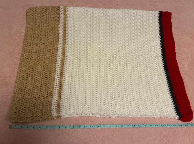 May include: A handmade crochet blanket with a color block design. The blanket features sections of tan, white, and red with a black stripe. The blanket is approximately 40 inches wide.