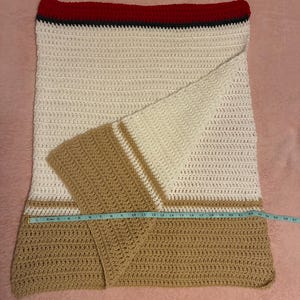 May include: A handmade crochet blanket with a cream, tan, and red color scheme. The blanket features a striped design with a red border and tan sections. The blanket is approximately 26 inches wide.