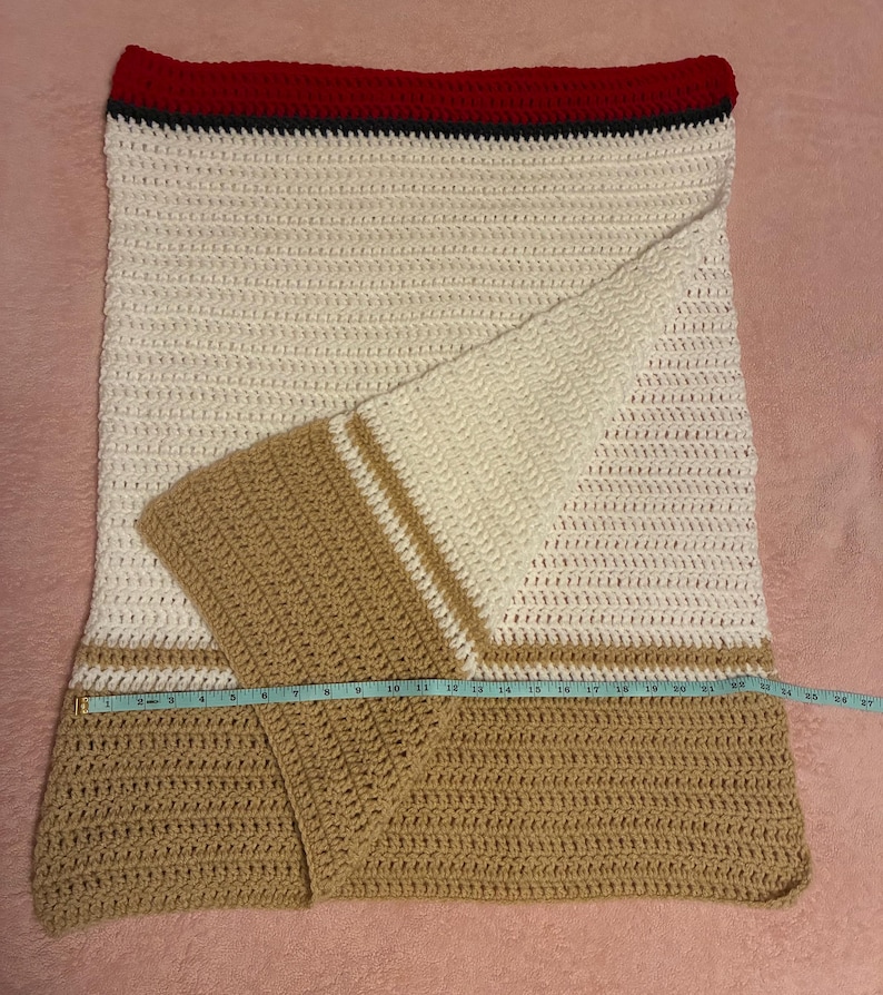 May include: A handmade crochet blanket with a cream, tan, and red color scheme. The blanket features a striped design with a red border and tan sections. The blanket is approximately 26 inches wide.