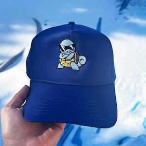 May include: A blue baseball cap with an embroidered image of a blue Squirtle wearing sunglasses. Squirtle is a Pokemon character.