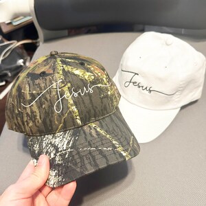 May include: Two baseball caps, one with a camouflage pattern and the other white. Both caps have the word "Jesus" embroidered on them in white cursive.
