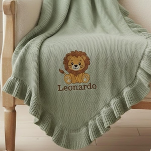 Può includere: Una coperta per bambini verde salvia con un bordo a balze. La coperta presenta un disegno di leone ricamato e il nome "Leonardo" in filo marrone. La coperta è drappeggiata su una sedia.
