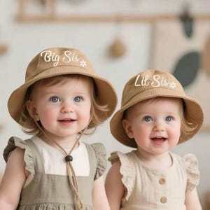 Puede incluir: Dos bebés con sombreros de cubo marrones con bordados blancos. El sombrero de la izquierda dice "Big Sis" y el sombrero de la derecha dice "Lil Sis".