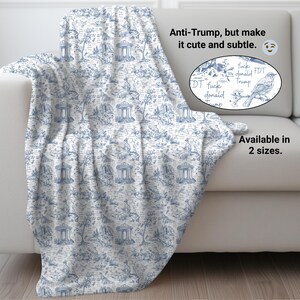May include: A white and blue throw blanket draped over a white sofa. The blanket features a toile pattern with trees, buildings, and figures. The text "Anti-Trump, but make it cute and subtle" is visible, along with the phrase "Available in 2 sizes."