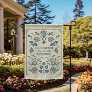 Op de afbeelding: Een tuinvaandel met een crèmekleurige achtergrond en een bloemenontwerp in blauw- en groentinten. De vlag bevat de tekst "Geradicaliseerd door elementaire fatsoenlijkheid."
