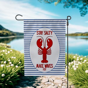 Puede incluir: Una bandera de jardín con un fondo a rayas azules y blancas. La bandera presenta un óvalo blanco con una ilustración de langosta roja y las palabras "STAY SALTY" y "MAKE WAVES" en texto rojo. La bandera está colgada de un soporte de metal negro.