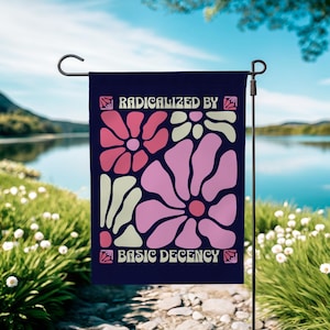 Peut inclure: Un drapeau de jardin bleu marine avec un motif floral rose et crème. Le drapeau porte le texte "RADICALIZED BY" en haut et "BASIC DECENCY" en bas, dans une police rétro.