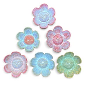 May include: Six ceramic flower-shaped dishes arranged in a triangular formation. Each dish features a central face with closed eyes and a textured, glazed finish in varying shades of pink, blue, green, and iridescent purple, yellow, and orange.