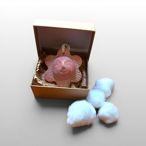 May include: A ceramic sun-shaped ornament with a smiling face, in a cardboard box with packing material. The ornament is a reddish-pink colour. Fluffy white cotton balls are next to the box.