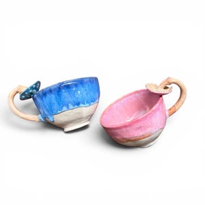 May include: Two ceramic mugs, one blue and one pink, with mushroom-shaped handles. The blue mug has a textured, dripped glaze, while the pink mug has a smooth finish. Both mugs have a cream-coloured base and are set against a white background.