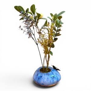 May include: A low, rounded ceramic vase with a glossy blue and white drip glaze finish. A small black and white bird figurine is attached to the rim. The vase contains a bouquet of artificial green eucalyptus stems and small yellow flowers.