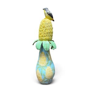 May include: A tall ceramic vase featuring a light blue base painted with yellow and green floral motifs. The top of the vase is adorned with a textured, three-dimensional yellow flower head, upon which sits a small, painted ceramic bird figure.