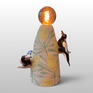 May include: A conical ceramic table lamp featuring a textured surface with a pale pink base, green leaves, and yellow oval patterns. Two black and white magpie figures are perched on small branches extending from the sides, topped with a round bulb.