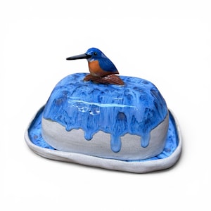 May include: A ceramic butter dish featuring a white base with a speckled finish and a lid adorned with a blue and orange kingfisher bird. The lid has a blue glaze that drips down the sides over the white speckled ceramic surface.