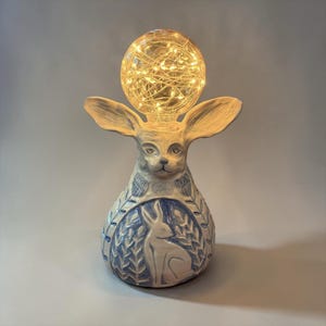 May include: A ceramic rabbit-shaped lamp with large ears, in blue and white. The base features a rabbit relief design. The light source is a globe filled with fairy lights, creating a warm glow. The lamp is a decorative item.