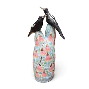 May include: A ceramic sculpture featuring two black birds with white wing markings perched atop a tall, bifurcated base. The base is decorated with a pattern of pink bell-shaped flowers and green leaves against a light blue dotted background.