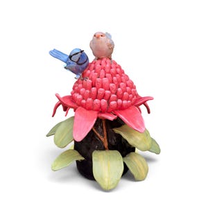 May include: A ceramic sculpture featuring a bright pink waratah flower with a textured centre, perched upon a dark brown stem with light green leaves. Two small birds, one blue and one light pink, are positioned on top of the flower petals.