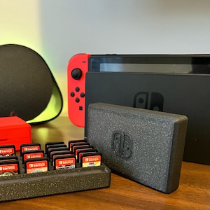Può includere: Una console Nintendo Switch nera con un controller Joy-Con rosso e una custodia nera. La console è appoggiata su un tavolo di legno con un porta cartucce di gioco nero e grigio e una custodia rossa. Il porta cartucce ha 12 slot per cartucce di gioco. Le cartucce sono etichettate "Nintendo Switch".