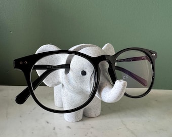 May include: Black-framed eyeglasses rest on a white and gray elephant figurine. The glasses have round lenses and the text "BEN & FRANK" is visible on the frame. The elephant is on a white surface against a green background.