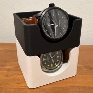 Könnte beinhalten: Ein zweistufiger Uhrenständer mit einer schwarzen oberen Stufe und einer weißen unteren Stufe. Die obere Stufe hält eine schwarze Tissot-Uhr mit einem braunen Lederband. Die untere Stufe hält eine silberne Uhr mit einem grünen Zifferblatt.