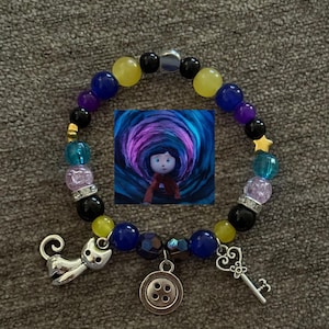 May include: A beaded bracelet featuring a central image of a girl in a tunnel. The bracelet includes charms of a cat, a key, and a button. Beads are in various colors including blue, yellow, purple, and black.