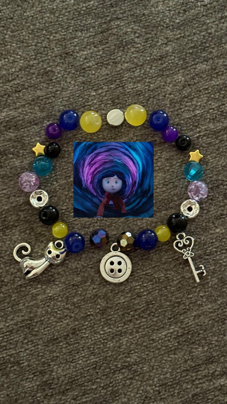 May include: A beaded bracelet featuring a central image of a character in a tunnel. The bracelet includes blue, yellow, purple, and clear beads, along with charms of a cat, a key, and a button. The bracelet is designed with a variety of colors and shapes.