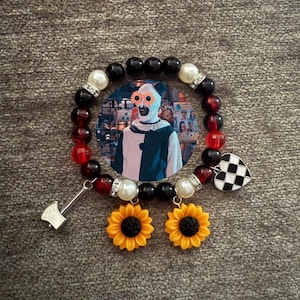 May include: A beaded bracelet with black, red, and white beads, a circular image of a clown, sunflower charms, and a checkered heart charm. A silver axe charm hangs from the bracelet.