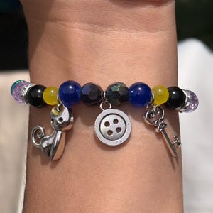 May include: A beaded bracelet with charms. The bracelet features a variety of beads in black, yellow, blue, and iridescent colors. Charms include a cat, a button, and a key. The bracelet is worn on a wrist.