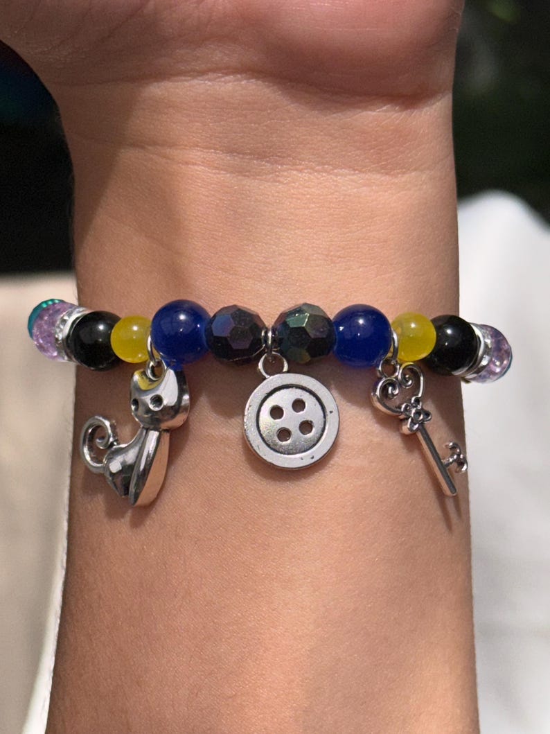 May include: A beaded bracelet with charms. The bracelet features a variety of beads in black, yellow, blue, and iridescent colors. Charms include a cat, a button, and a key. The bracelet is worn on a wrist.