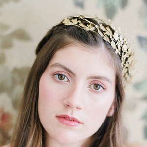 May include: A woman with long brown hair wearing a gold leaf crown and a white lace top.