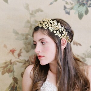 May include: A woman wearing a white lace dress and a gold leaf crown sits in front of a floral painting.