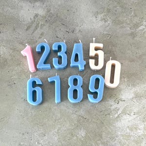 以下が含まれることがあります： さまざまな色の数字型キャンドルのコレクション。数字の1、6、7、8、9は青色で、5と0はオフホワイトです。数字の1はピンク色です。キャンドルは、質感のある灰色の表面に配置されています。