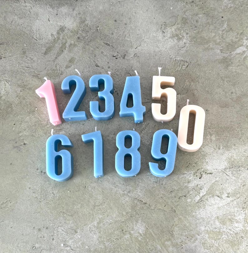 以下が含まれることがあります： さまざまな色の数字型キャンドルのコレクション。数字の1、6、7、8、9は青色で、5と0はオフホワイトです。数字の1はピンク色です。キャンドルは、質感のある灰色の表面に配置されています。