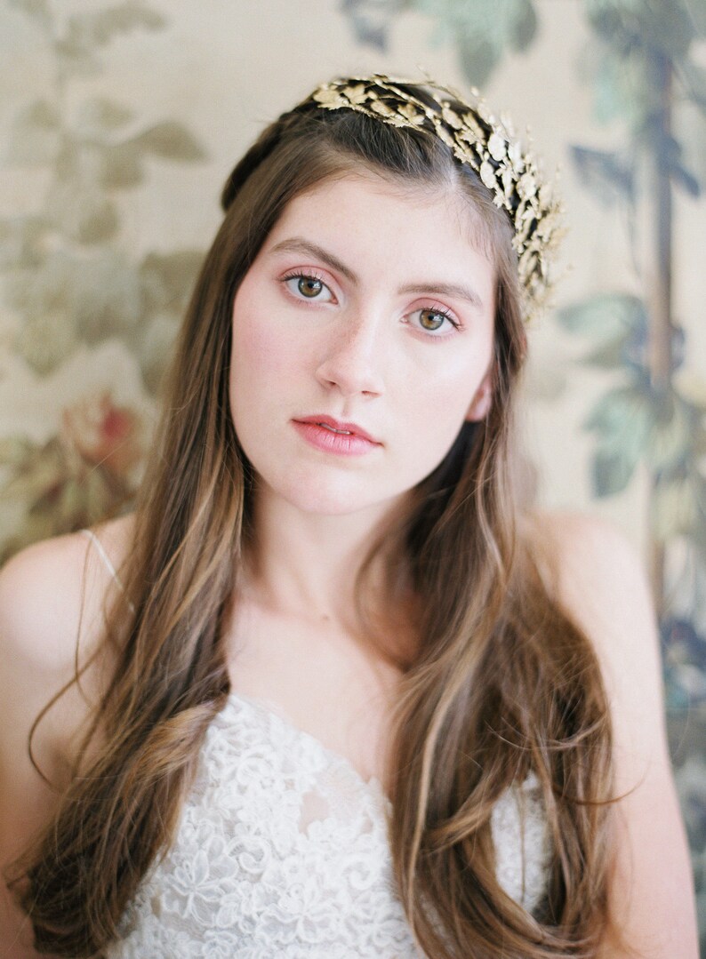 May include: A woman with long brown hair wearing a gold leaf crown and a white lace top.