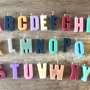 以下が含まれることがあります： 木製の表面に配置された、カラフルな文字型のキャンドルのコレクション。キャンドルはピンク、青、黄色、緑など、さまざまな色があります。各キャンドルには芯が付いています。AからZまでの文字が表現されています。