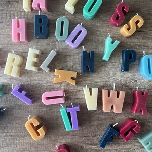 以下が含まれることがあります： ピンク、青、緑、黄色など、さまざまな色合いのカラフルな文字型キャンドルのコレクション。各キャンドルには芯があり、木製の表面に配置されています。キャンドルは単語やフレーズを綴るように設計されています。
