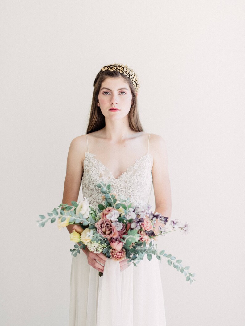 May include: A bride wearing a white lace wedding dress with a gold leaf crown and holding a bouquet of pink, purple, and yellow flowers.