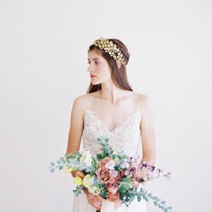 May include: A bride wearing a white lace dress and a gold leaf crown holds a bouquet of pink, purple, and yellow flowers.